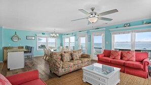 A spacious living room with a large window overlooking the sea, a ceiling fan, and a dining area with a table and chairs. - The Perfect Oceanfront Vacation w/ Private Pool! (Holden Beach)