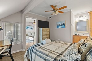 Un dormitorio con cama, ventilador de techo, televisor, cómoda, escritorio y ventana con persianas.