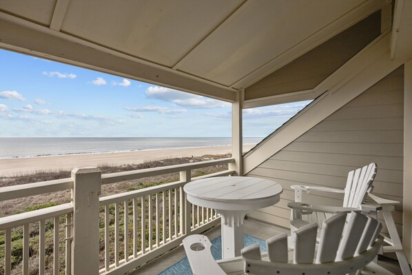 A covered balcony with a view of the beach, white Adirondack chairs, and a small round table.