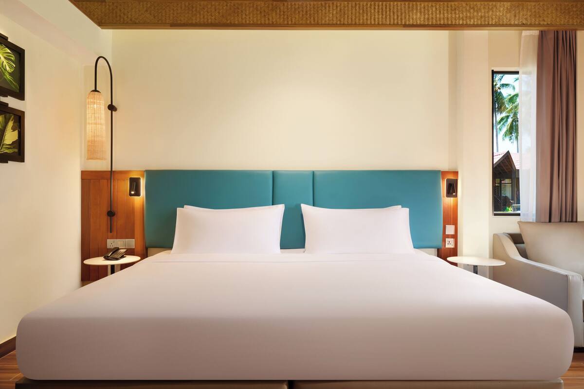 Habitación de hotel con una cama grande, una mesita de noche de madera, una pequeña mesa, una silla y vistas a palmeras.