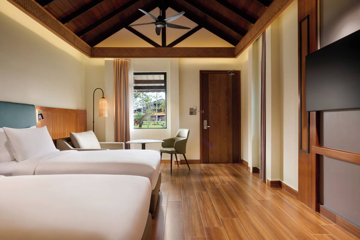 Habitación de hotel con dos camas, ventilador de techo, suelo de madera, ventana con cortinas y una puerta.