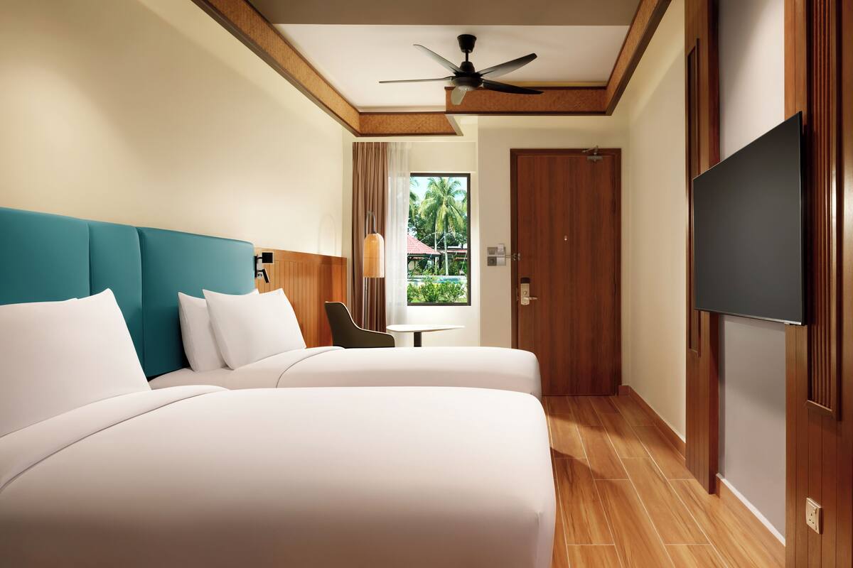 Habitación de hotel con dos camas, ventilador de techo, televisor, una mesita y ventana con vistas a palmeras.