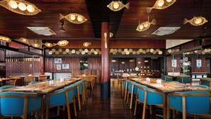 A restaurant interior with wooden tables and blue chairs, fish-shaped hanging lights, and a central pillar. - Hilton Garden Inn Kota Kinabalu Tuaran (Tuaran)