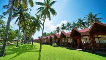 A row of bungalow-style accommodations with red roofs, surrounded by lush green lawns and tall palm trees under a clear blue sky.