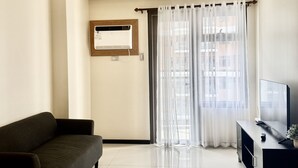 A modern living room with a sofa, a flat-screen TV, and a large window with sheer curtains.