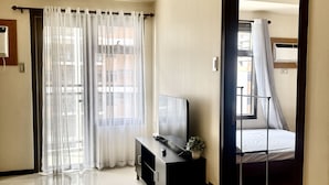 A hotel room with a large bed, a TV on a stand, a small table with a plant, and sheer curtains on the windows.