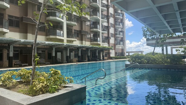 A swimming pool with a tiled floor, surrounded by a building with multiple balconies and a covered area with seating.