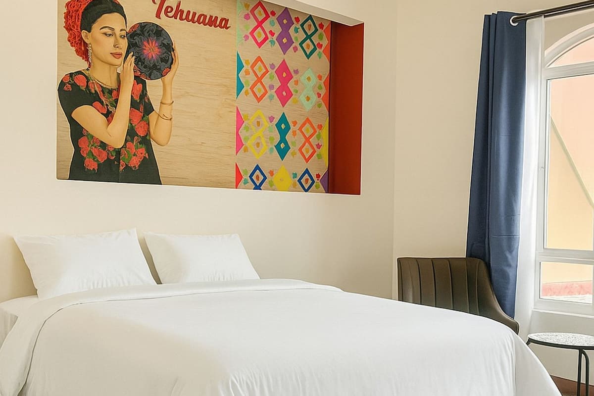 A hotel room with a bed, a chair, a window with curtains, and a wall with a colorful geometric pattern and a poster.