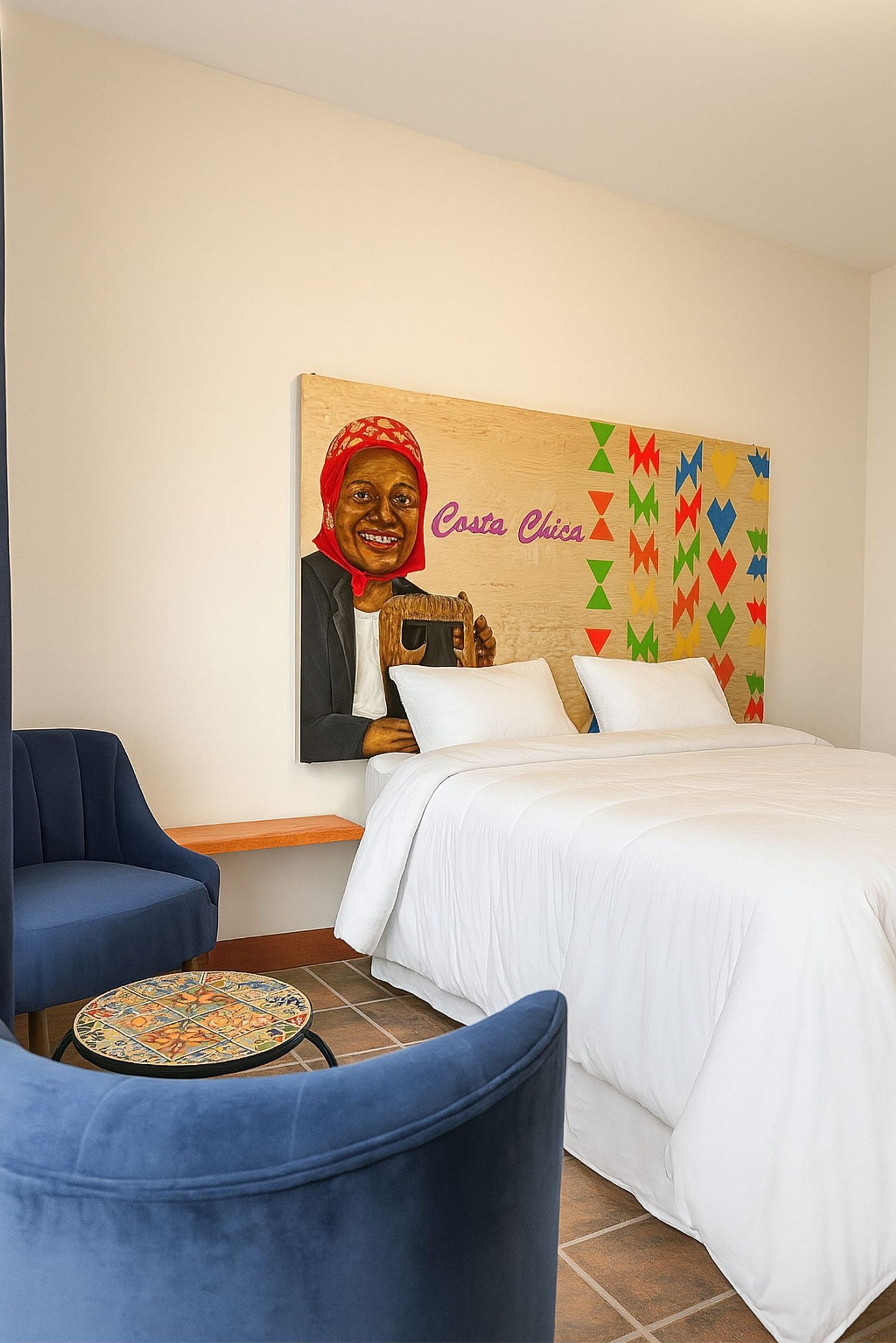 Une chambre d’hôtel avec un lit, deux fauteuils bleus, une petite table ronde et un élément de décoration murale aux couleurs vives.