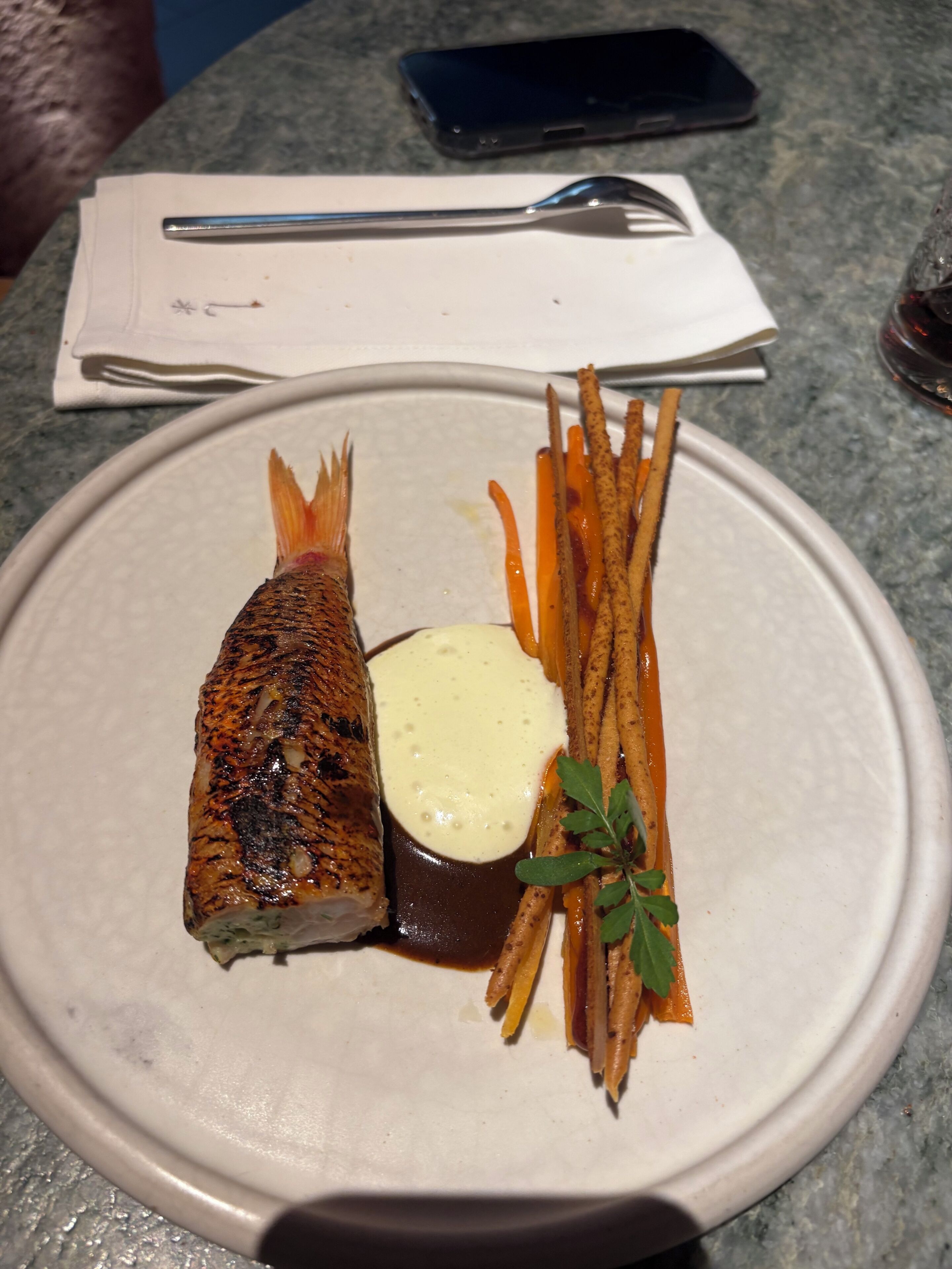 A plate with grilled fish, fries, and a sauce, with a napkin and a phone on the table.