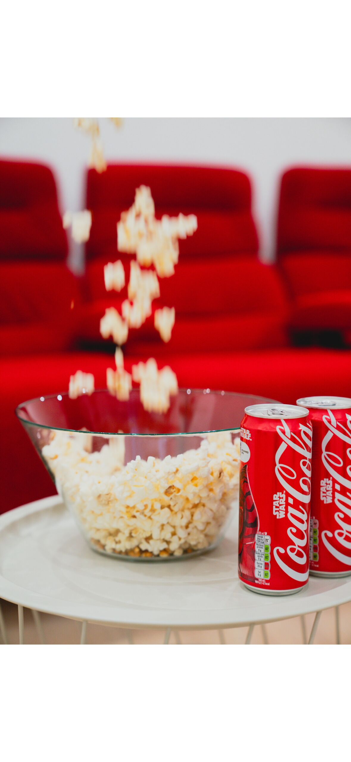 A glass bowl filled with popcorn and two cans of Coca-Cola on a white table.