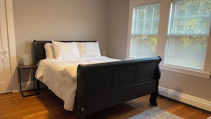 Une chambre à coucher avec un lit, une table de chevet, une fenêtre avec des stores et un plancher en bois.