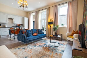 Ein modernes Wohnzimmer mit einem blauen Sofa, einem Couchtisch und einem Fernseher. Im Hintergrund befindet sich eine Küche mit weißen Schränken und ein Essbereich mit orangefarbenen Stühlen.