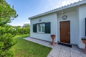 Ein eingeschossiges Gebäude mit weißer Fassade, einer braunen Tür und grünen Fensterläden. Vor dem Haus befindet sich ein kleiner Garten mit Rasen und einem Blumentopf.
