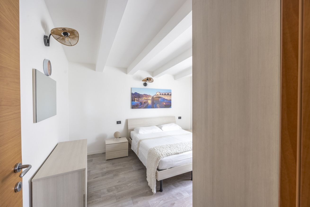 A bedroom with a bed, bedside tables, a wall-mounted lamp, and a painting of a cityscape.