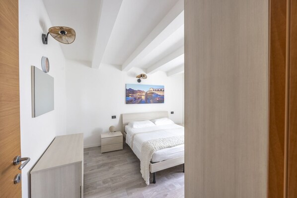 A bedroom with a bed, bedside tables, a wall-mounted lamp, and a painting of a cityscape.