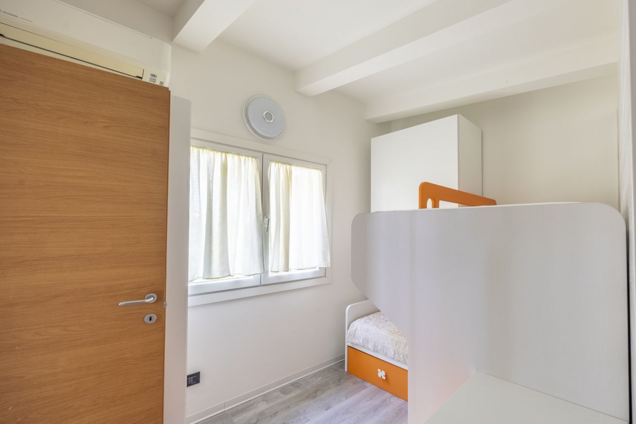 Ein Schlafzimmer mit Holztür, einem fenster mit Vorhängen und einem Etagenbett, wobei die untere Etage orange ist, sowie einem weißen Kopfteil.