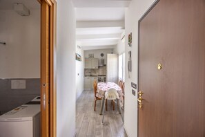 A narrow hallway leading to a dining area with a table and chairs, a kitchen in the background, and a wooden door on the right.