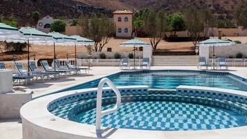 Une piscine dotée de chaises longues et de parasols, entourée d’un espace paysager avec une chaîne de montagnes en toile de fond.