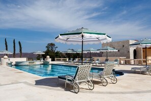 Un área de piscina con sillas de descanso y sombrillas, un muro de piedra y cielo despejado.