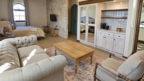 Un salón amplio con un sofá grande, una mesa de centro de madera y una zona de cocina con armarios blancos y un salpicadero de azulejos.