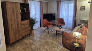 A living room with a wooden cabinet, two armchairs, a small round table, a TV, and a radiator.