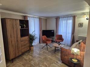 A living room with a wooden cabinet, two armchairs, a small round table, a TV, and a radiator.