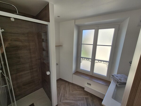 A small bathroom with a shower, a window with frosted glass, and a bench with towels.