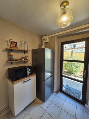 Eine kleine Küche mit einem Edelstahlkühlschrank, einer Mikrowelle und einem Holzkasten. Ein Regal mit Glasflaschen und ein Bilderrahmen sind ebenfalls vorhanden. Eine Tür führt auf einen Balkon mit Blick ins Grüne.