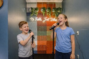 Deux enfants tenant des microphones devant un mur orné d’un motif coloré et texturé, avec un panneau néon sur lequel on peut lire : « Pas de basses, juste de bonnes ondes. »