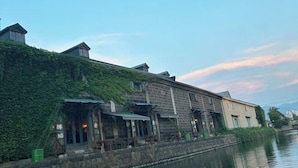 A riverside building with a stone facade, a covered outdoor seating area, and a small structure with a sign. - Minami Otaru House Sakaimachi (Otaru)