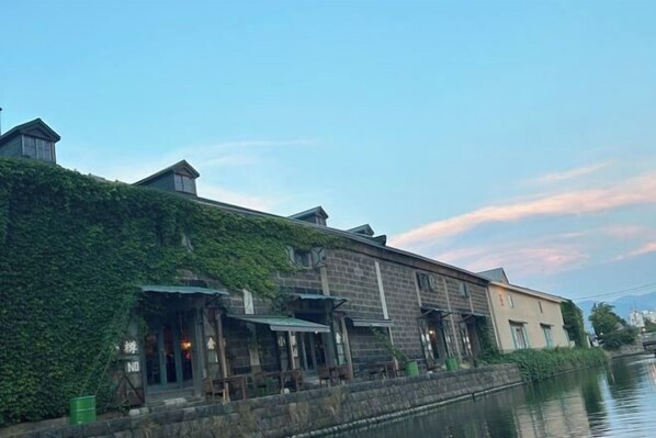 A riverside building with a stone facade, a covered outdoor seating area, and a small structure with a sign. - Minami Otaru House Sakaimachi (Otaru)