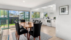 A modern kitchen with white cabinets, a built-in oven, and a coffee maker. There is a dining area with a wooden table and black chairs.