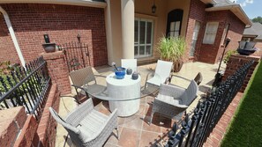 An outdoor patio with a glass table surrounded by wicker chairs, a potted plant, and a brick wall. - Courtyard Haven Stylish 3BR Retreat (Prairieville)
