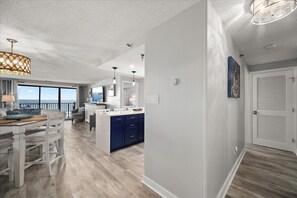 A modern kitchen with a central island, a dining area with a view, and a hallway leading to a door. - Sandy Square – Direct Oceanfront Condo  3BR • 2BA • Sleeps 11 (Ocean City)