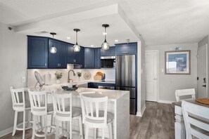 A modern kitchen with dark blue cabinets, stainless steel appliances, and a central island with white chairs. - Sandy Square – Direct Oceanfront Condo 3BR • 2BA • Sleeps 11 (Ocean City)