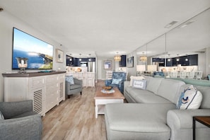 A modern living room with a large flat-screen TV, a grey sofa, a coffee table, and a kitchen area in the background. - Sandy Square – Direct Oceanfront Condo 3BR • 2BA • Sleeps 11 (Ocean City)