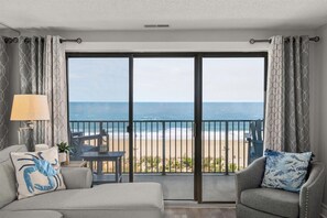 A balcony with a view of the beach, a sofa, a chair, and a table with a lamp and a plant. - Sandy Square – Direct Oceanfront Condo 3BR • 2BA • Sleeps 11 (Ocean City)