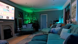Ein Wohnzimmer mit einem im grünen Licht leuchtenden Fernseher, einem Kamin, einer Couch und einer Topfpflanze.