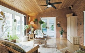 A spacious living room with a wooden ceiling, a ceiling fan, a large window, and a brick wall.