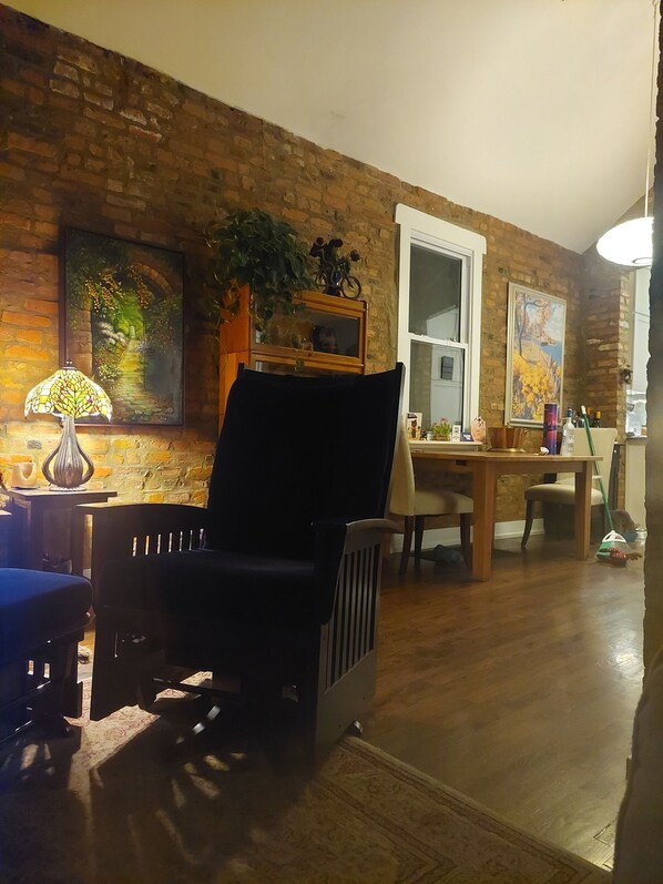 Una sala de estar con pared de ladrillo, una silla negra, una mesa con sillas y una ventana.