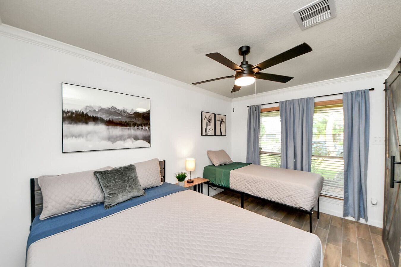 A bedroom with two beds, a ceiling fan, a window with curtains, and a wall-mounted picture.