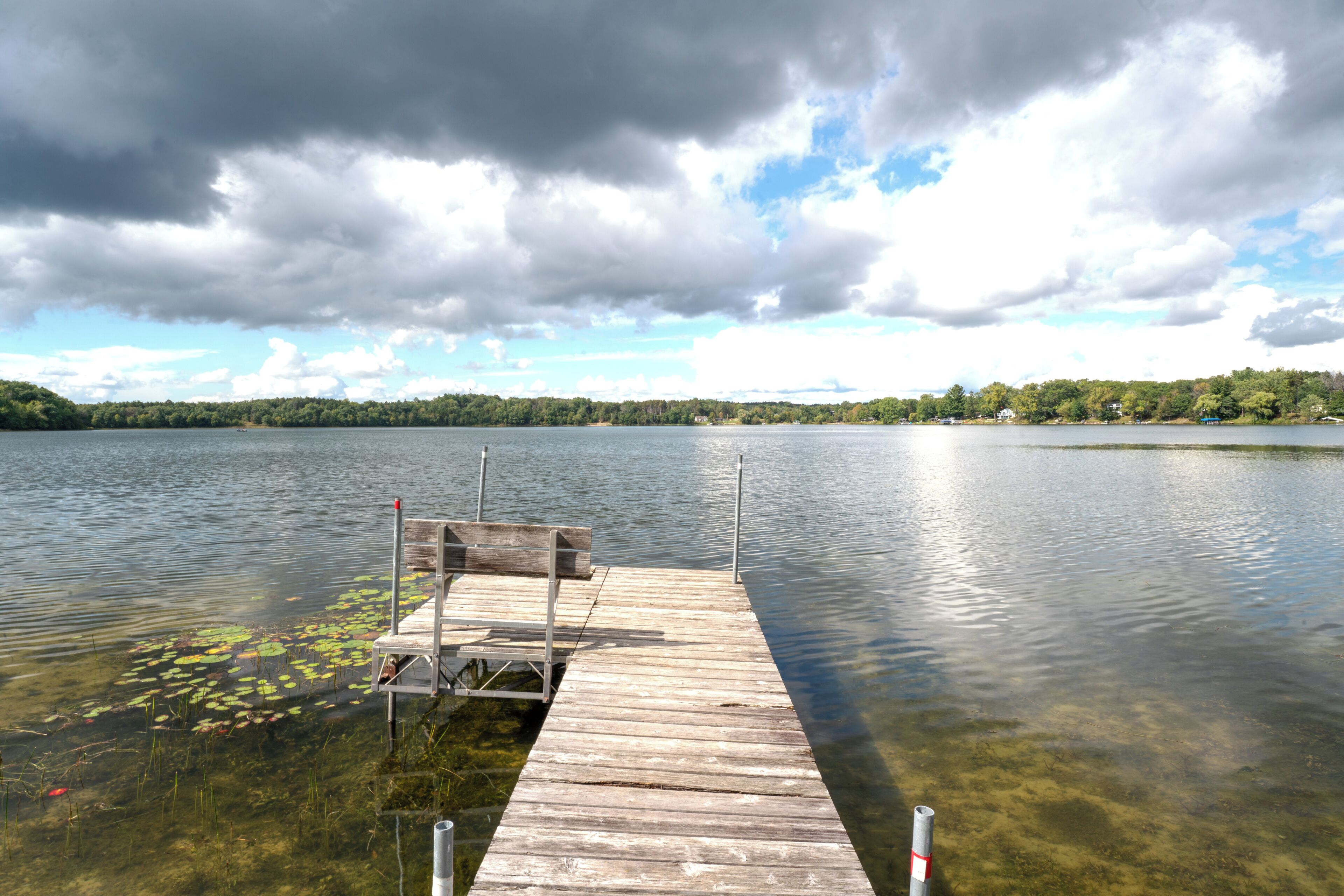 A wooden dock extending into a calm lake with clear water and lily pads.