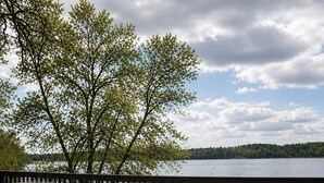 Une terrasse en bois avec une table et des chaises, donnant sur un lac bordé d’arbres et surplombant un ciel nuageux.