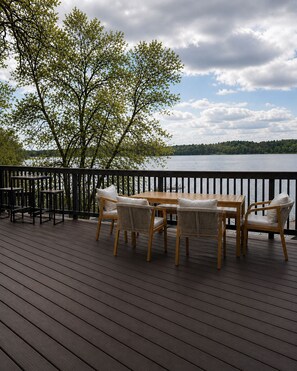 Terraza de madera con una mesa y sillas, con vistas a un lago rodeado de árboles y un cielo nublado.