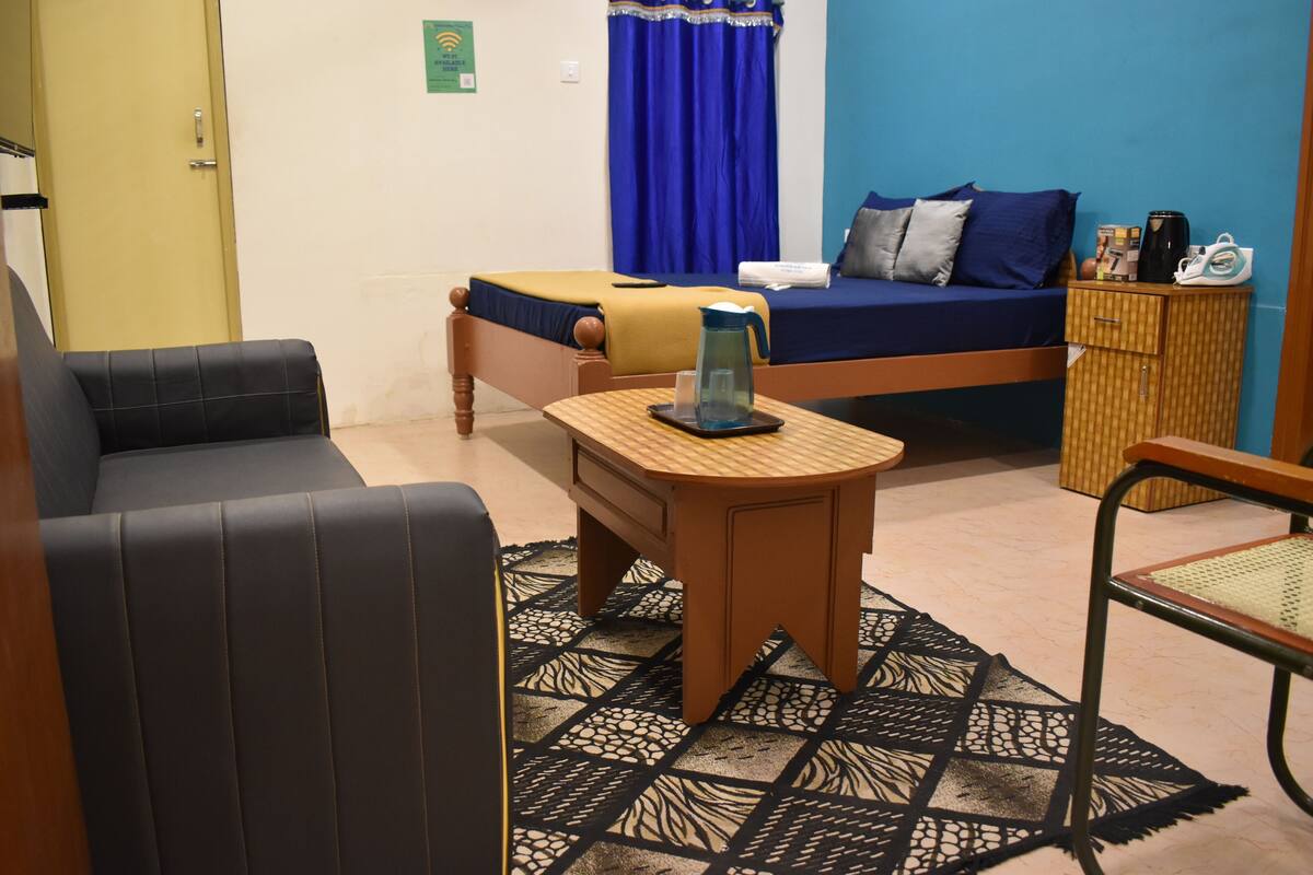 A room with a bed, a sofa, a small table with a kettle, and a chair.