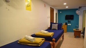 Habitación con dos camas, cada una con cubrecama azul y una toalla enrollada. Se ve una mesita de noche de madera con una lámpara y una pequeña mesa con un ventilador. Un televisor está montado en la pared, y se distingue una puerta al fondo.
