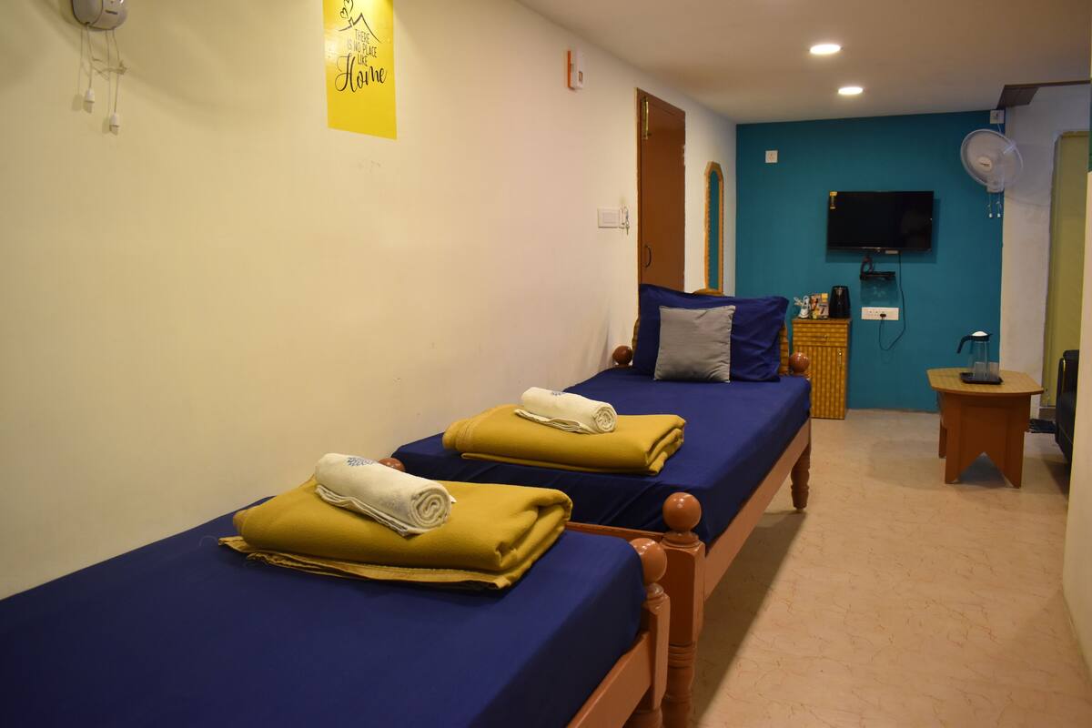 A room with two beds, each with a blue bedspread and a rolled towel. There is a wooden nightstand with a lamp and a small table with a fan. A television is mounted on the wall, and a door is visible in the background.