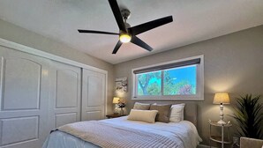 Et soveværelse med seng, ventilator, vindue, natbord, lampe og en potteplante.