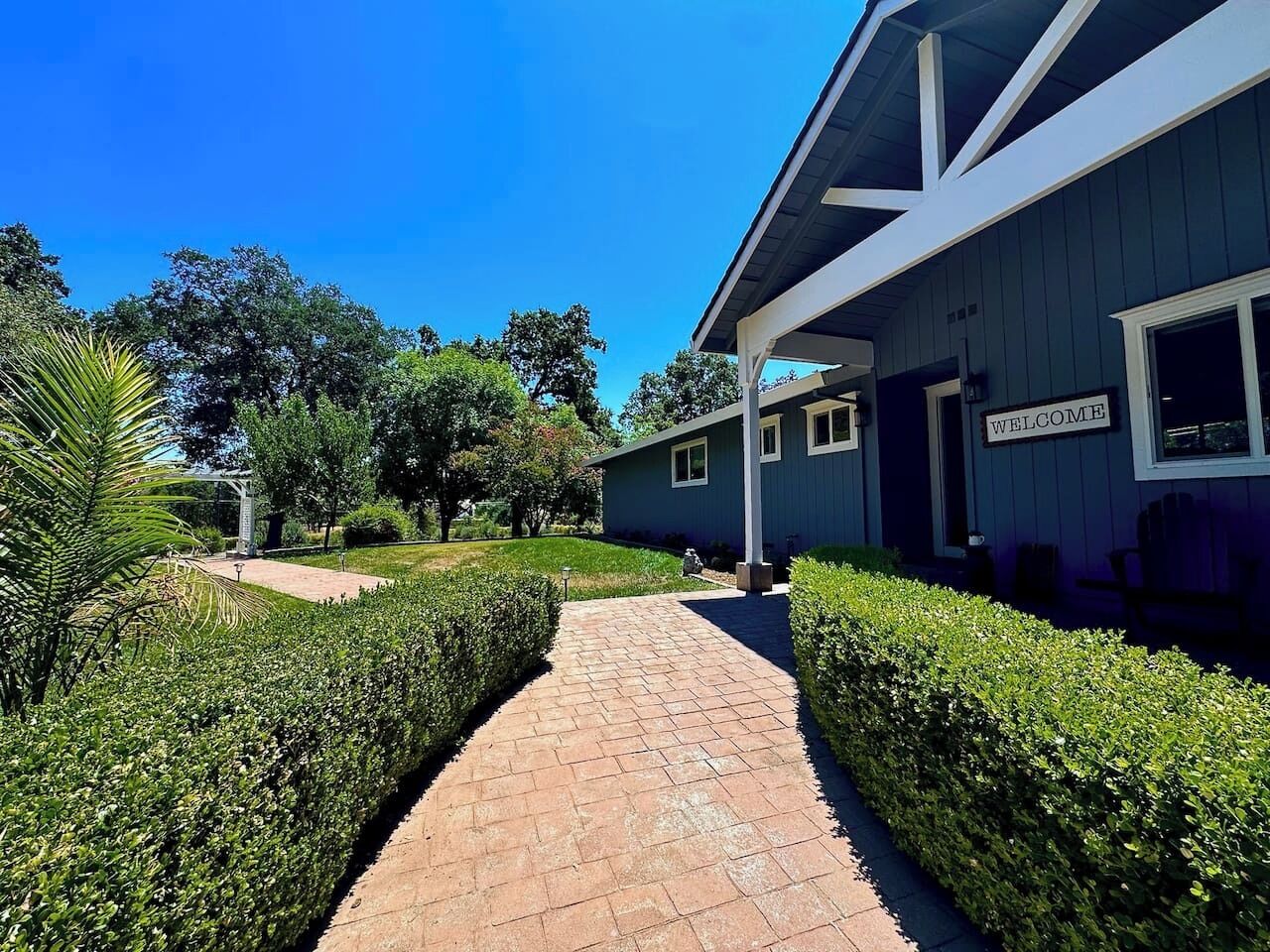 A pathway leading to a building with a 'WELCOME' sign, surrounded by well-maintained hedges and trees.